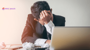 A person appears stressed while sitting at a desk, resting their head in their hand, with an open laptop and scattered crumpled paper balls