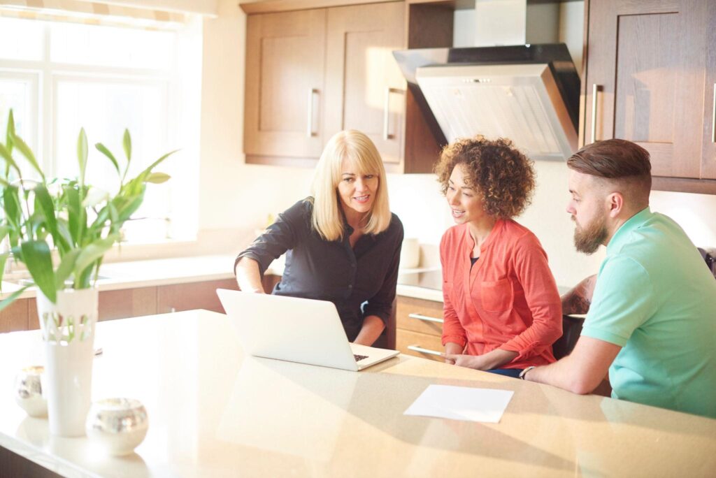 Mortgage adviser meeting with a first-time buyer couple at home, discussing mortgage options and affordability using a laptop.