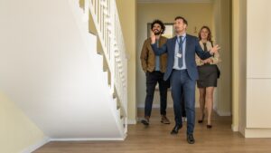Estate agent showing a couple around a modern property during a house viewing, walking through a bright hallway with wooden flooring and staircase.